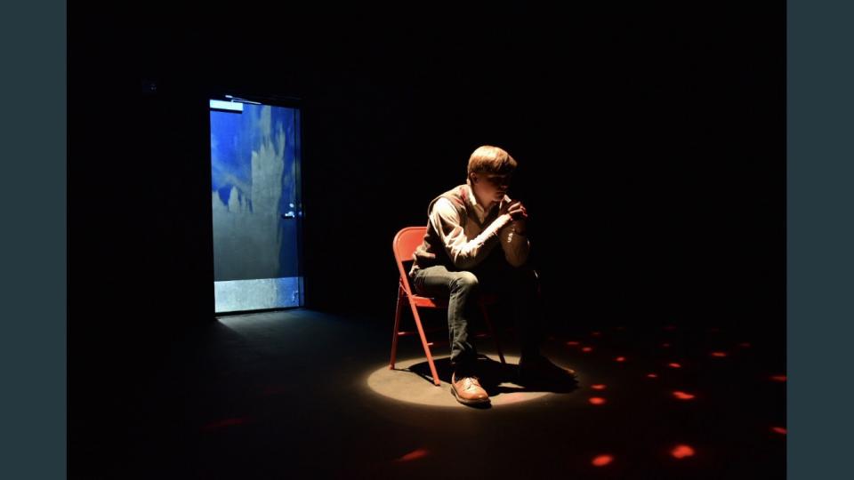 Student in spotlight sitting thoughtfully in folding chair on dark stage.